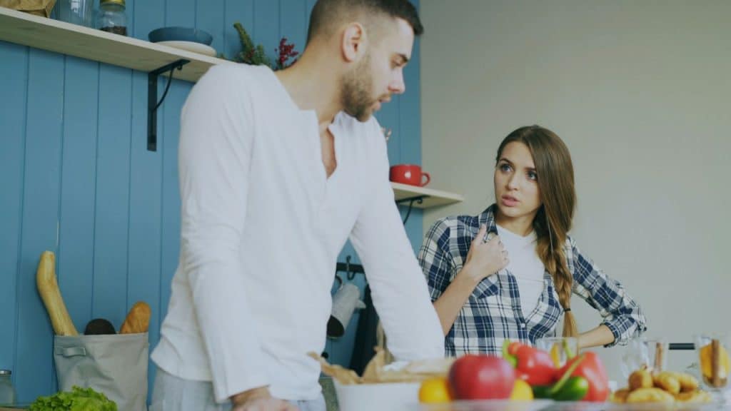 A man and woman in a kitchen look at each other with serious expressions near food.