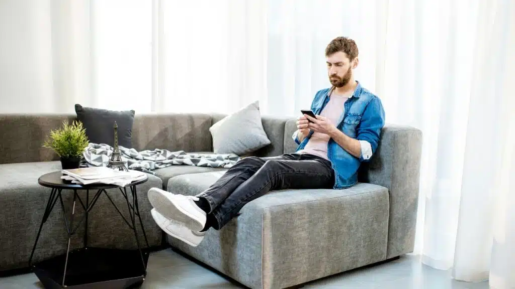 A bearded man sits on a grey couch in a bright room, looking at a smartphone.
