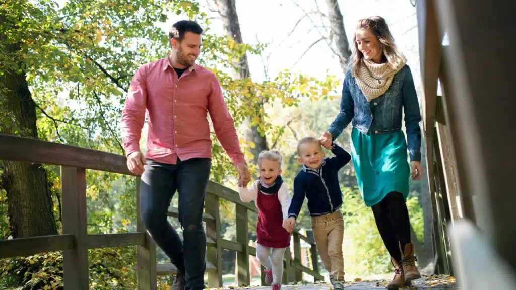 A man and woman walk outdoors on a wooden bridge holding hands with two children.