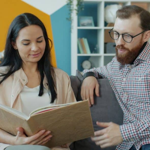 A woman and a bearded man sit on a gray sofa while looking at a book.