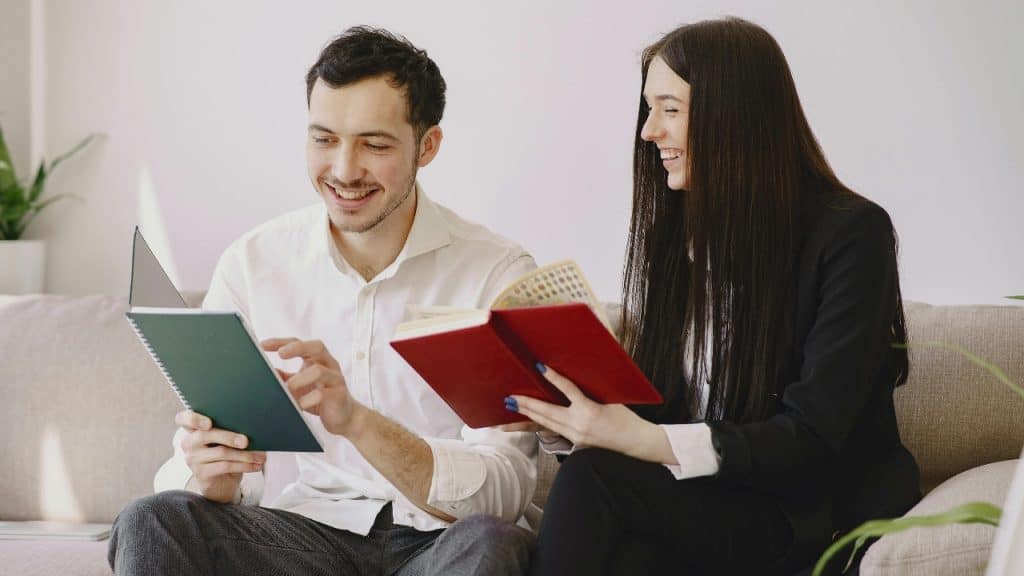 A man and woman sit on a couch laughing while holding open notebooks and pens.