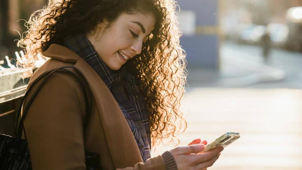 A woman smiling while looking at her phone outdoors.