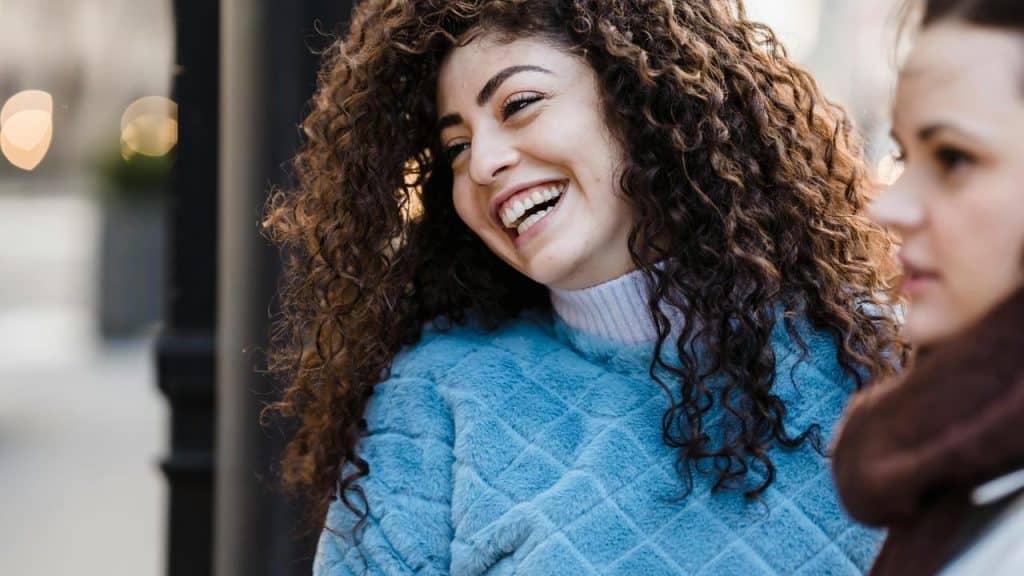 A woman with curly hair laughs while talking outdoors.