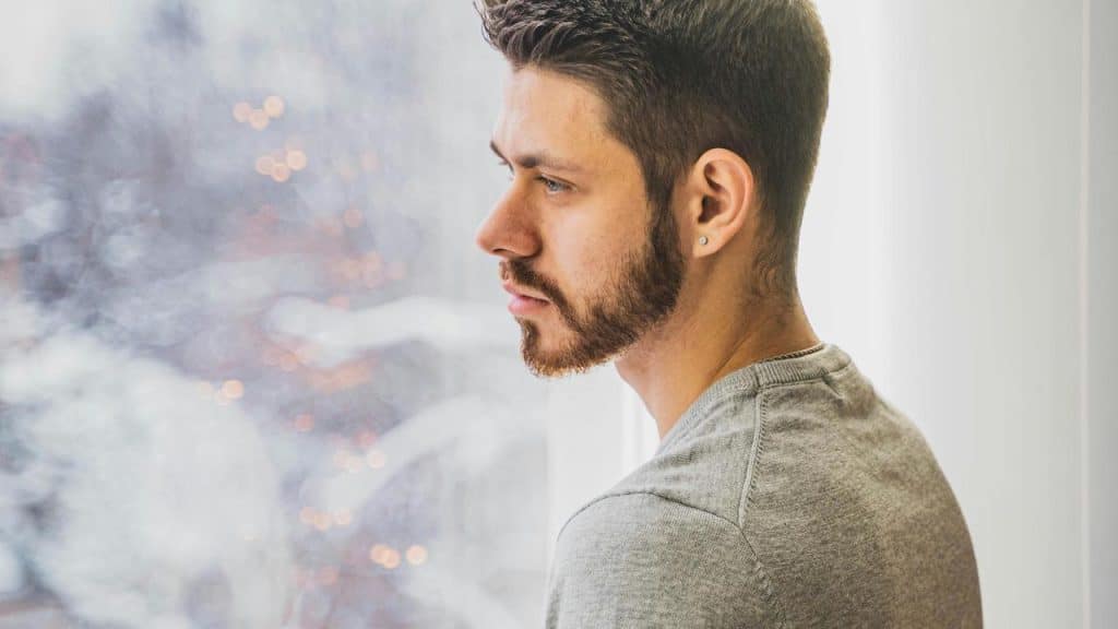 A man with a trimmed beard looking pensively out a bright window.