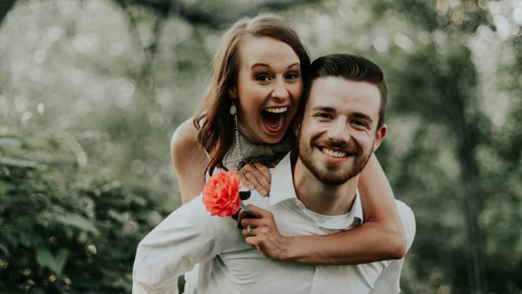 A cheerful woman holding a rose is riding on her husband’s back.