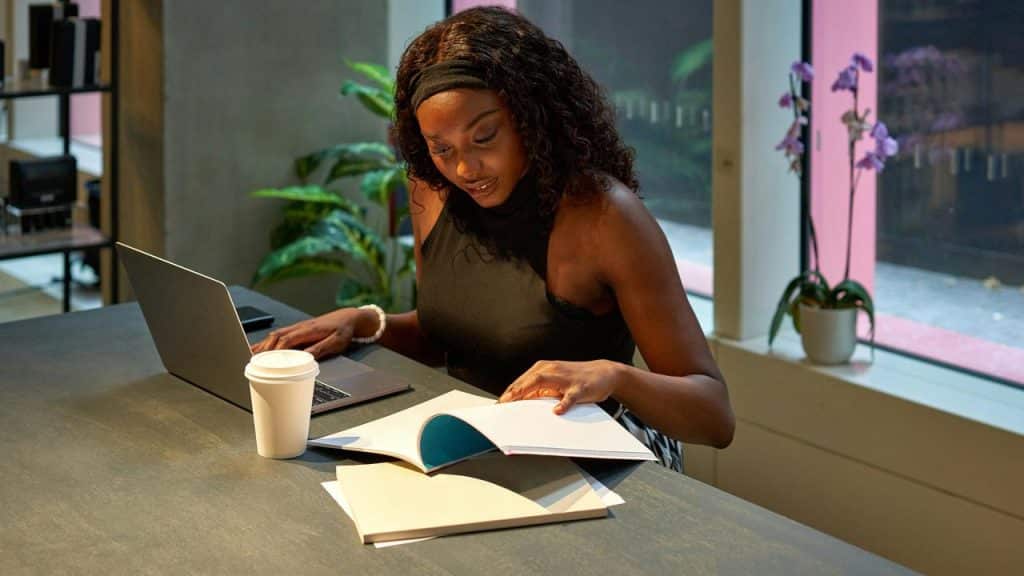 A woman reading a book at a table with a laptop and coffee in a modern workspace.