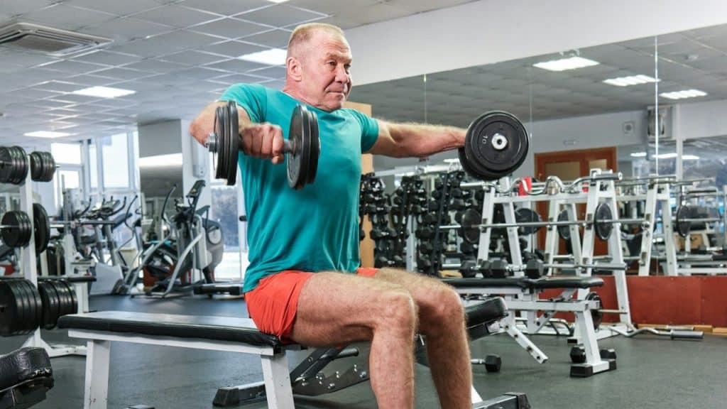 An older man in a teal shirt performs lateral raises with dumbbells in a gym.