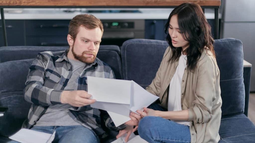 A man and woman sitting on a dark sofa holding and looking at white papers.