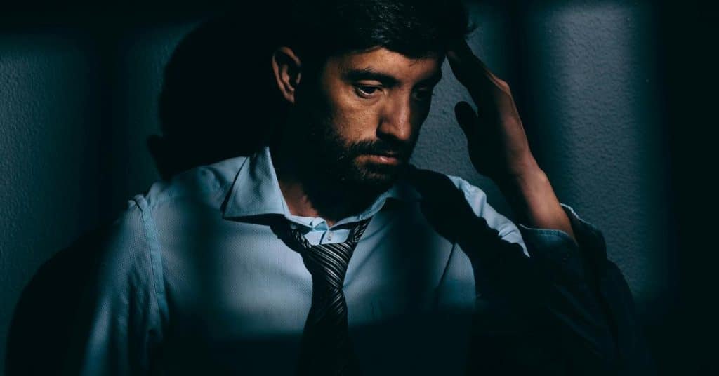 A man in a loosened tie holding his head in dim light.