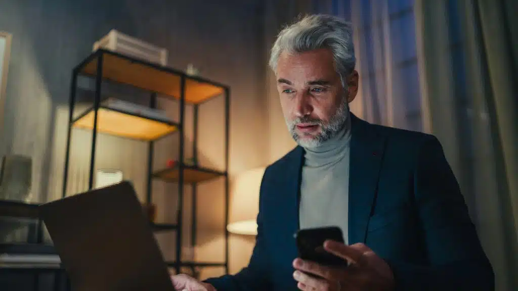 A middle-aged man with gray hair looks at a laptop while holding a smartphone.