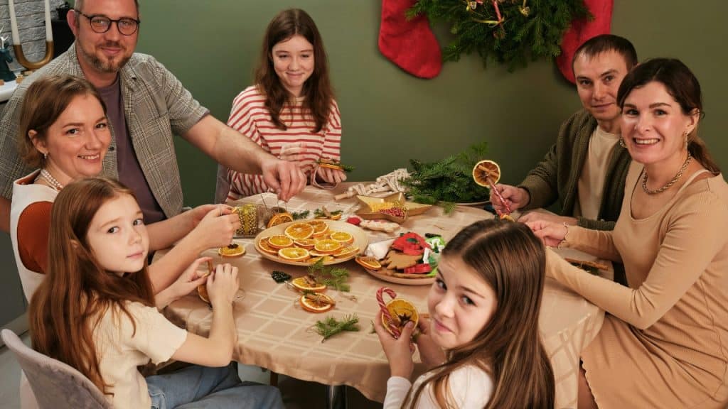 Six people of various ages sit around a table decorating orange slices and cinnamon sticks.