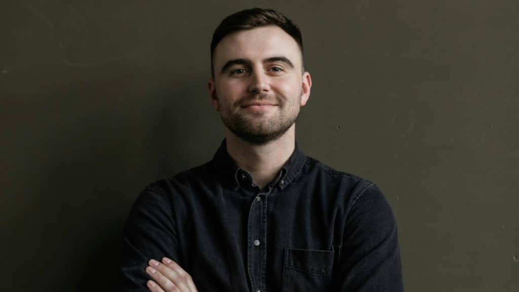 A man with short dark hair and a beard smiles while standing with his arms crossed.