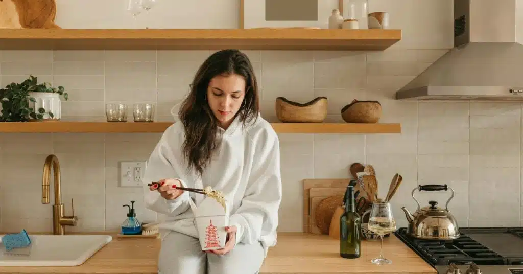 A person sits on a kitchen counter eating noodles from a takeout box.