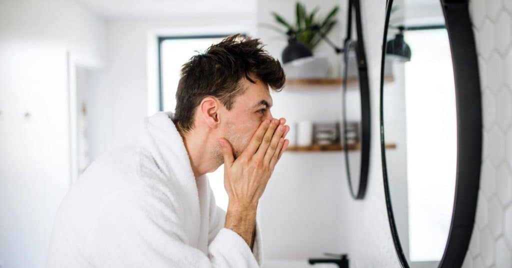 A man in a bathrobe washing his face while looking in a bathroom mirror.