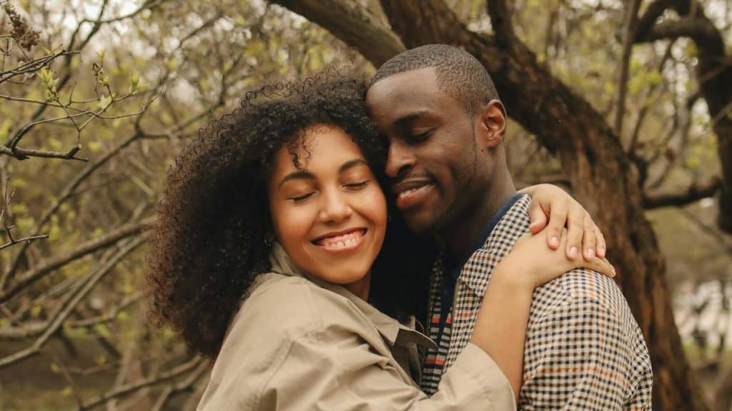 A smiling couple hugging closely outdoors among trees.