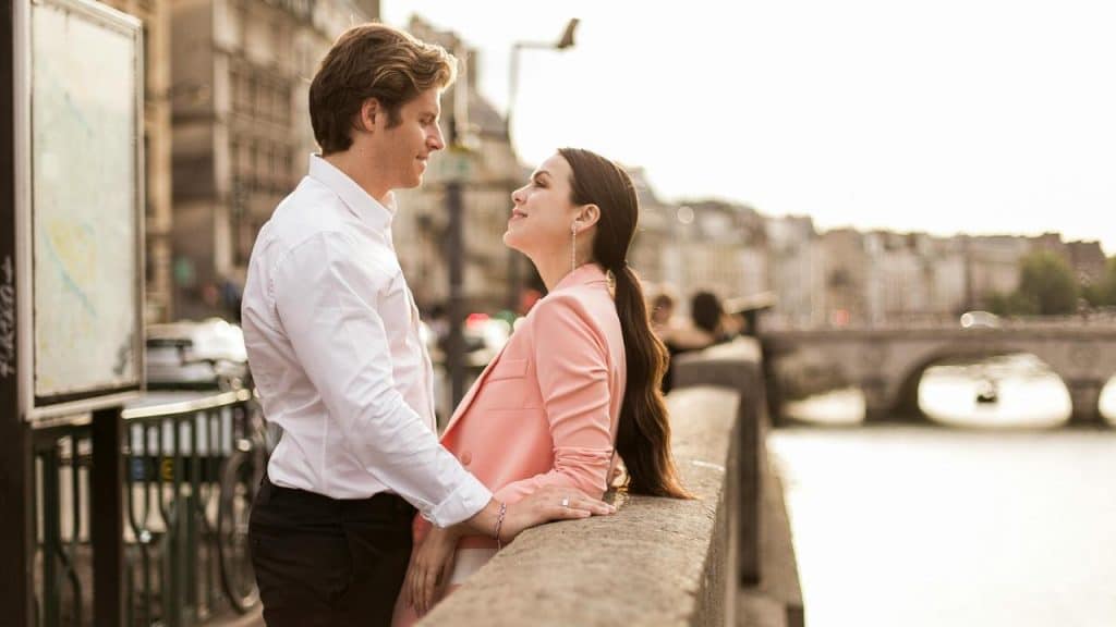 A couple standing close together by a river, smiling at each other in soft sunlight.