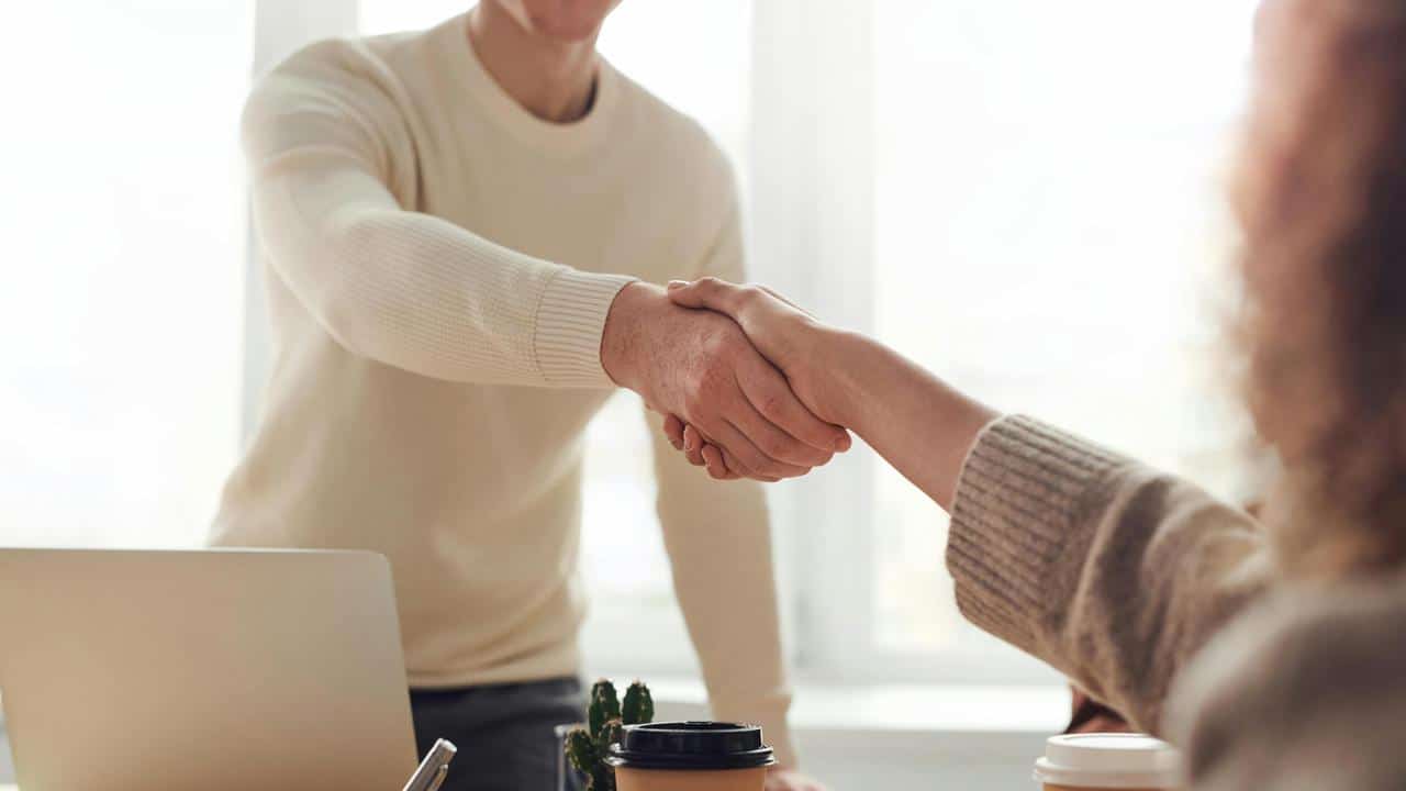 A man and woman shaking hands across a desk in a bright office.
