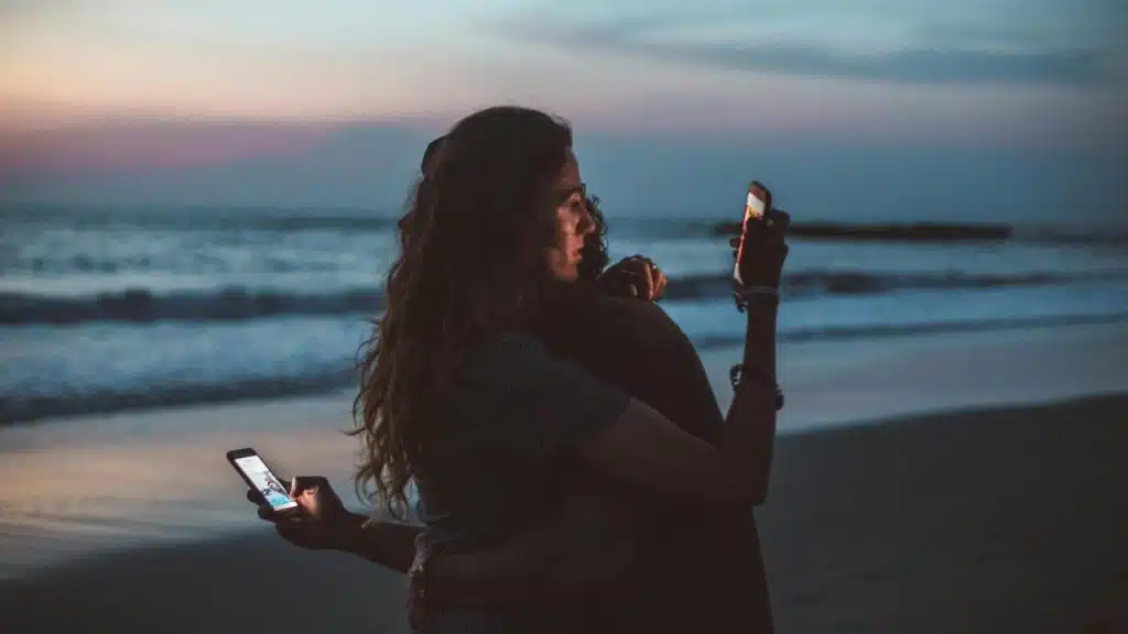 A couple sitting on a beach at dusk while looking at their phones.