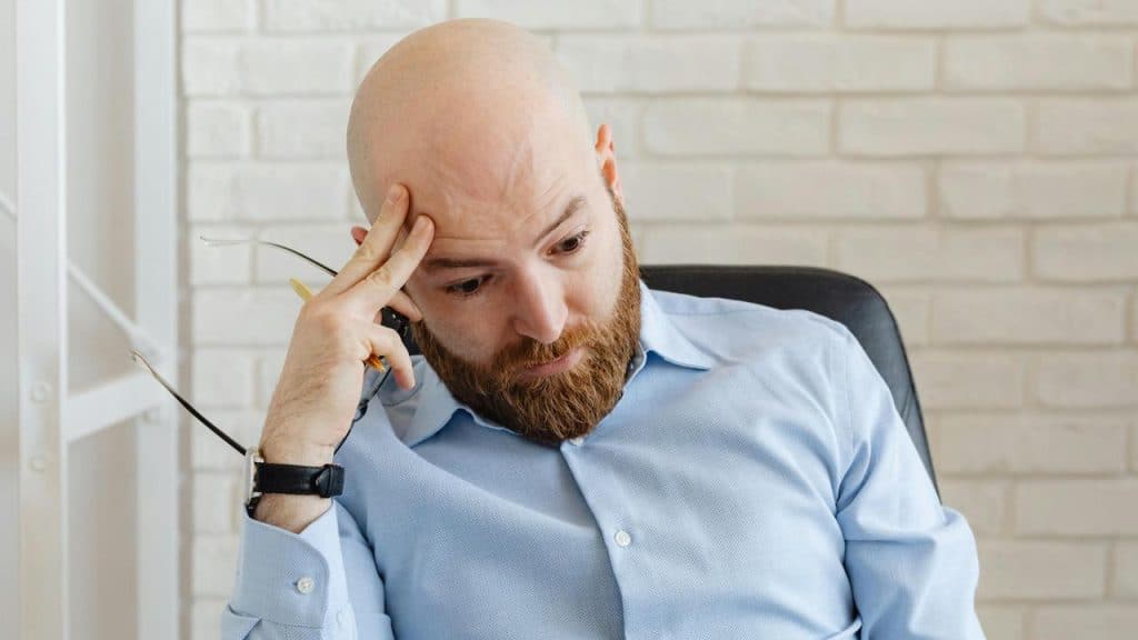 A man holding his head with one hand and glasses in the other looking down.