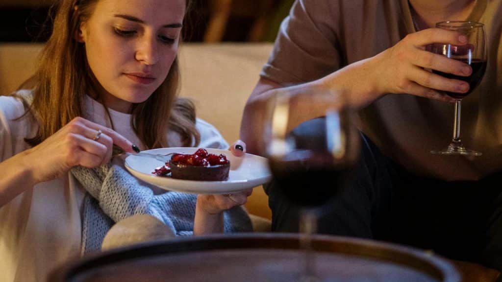 A woman eating strawberry cake while another person next to them holds a glass of wine.