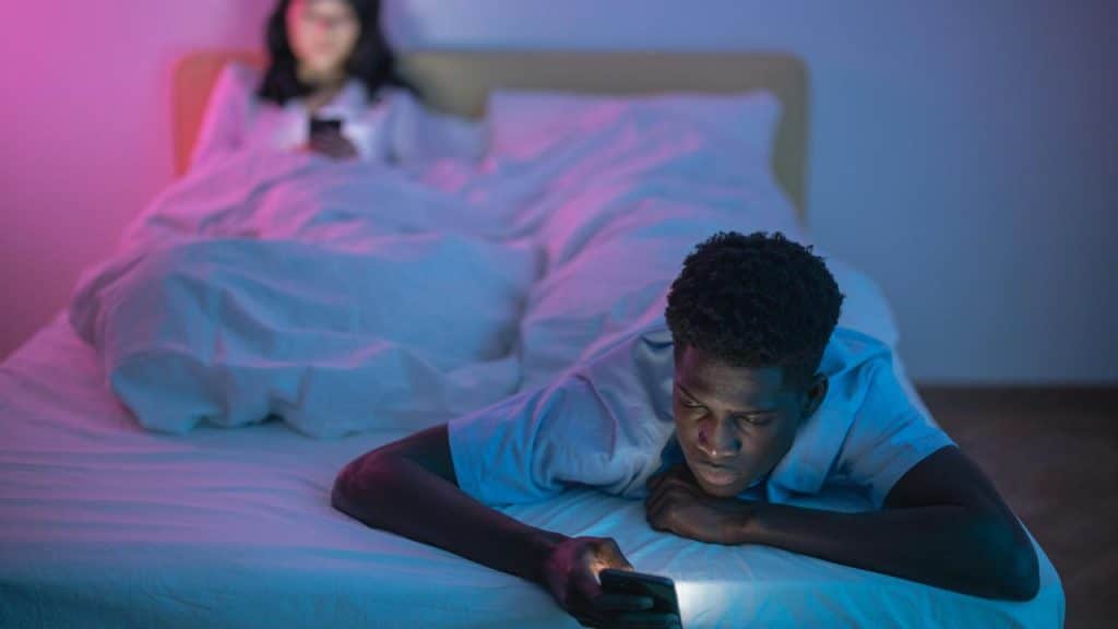 A man lying on a bed using his phone while another person sits in the background.