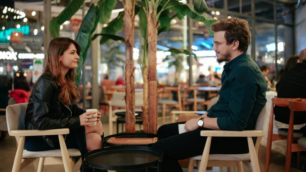 A man and woman sit at a small table in a cafe holding coffee mugs.