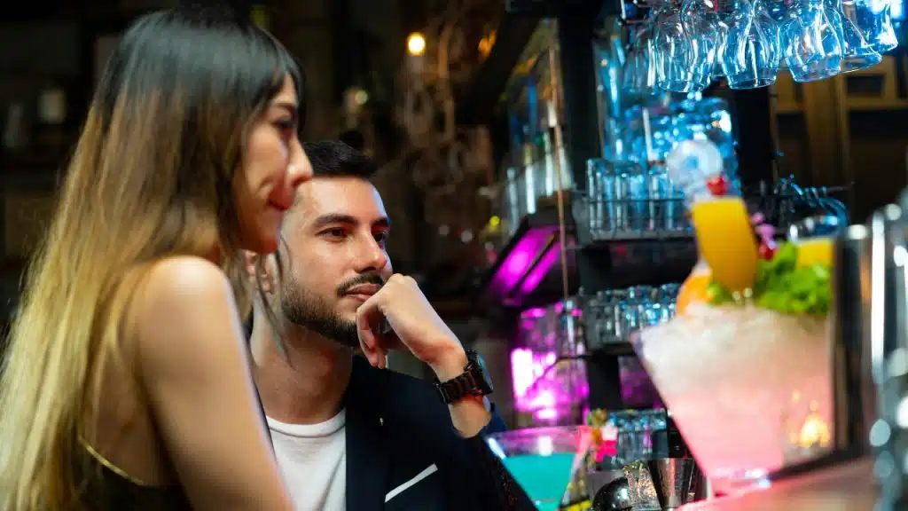 A man and woman sit at a bar with colorful cocktails and glasses in the background.