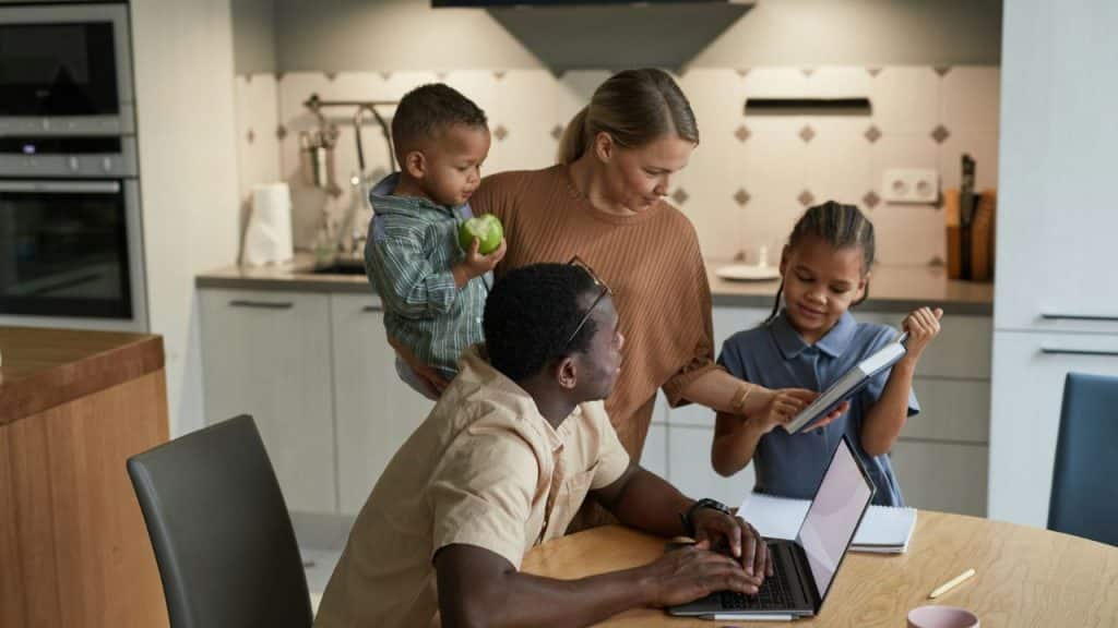 A man works on a laptop at a table while a woman and two children stand nearby.