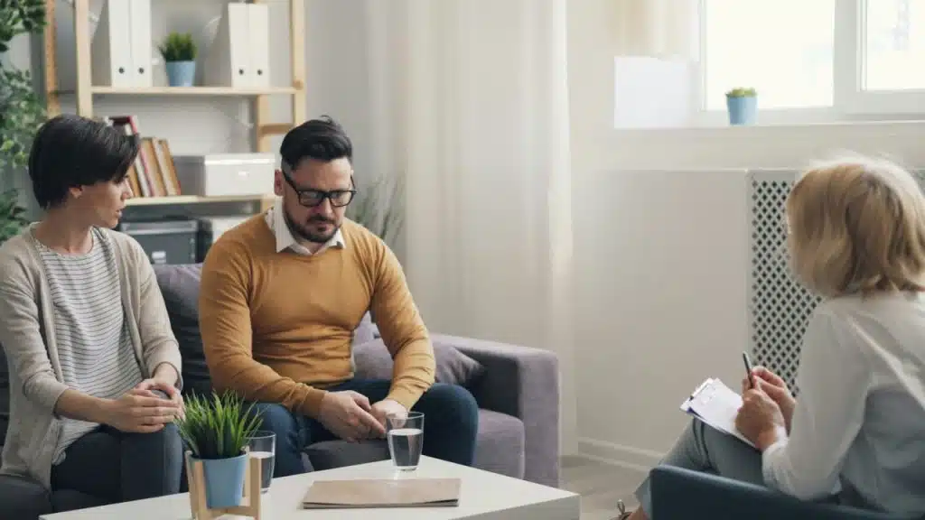 A man and woman sit on a couch facing a person holding a clipboard.