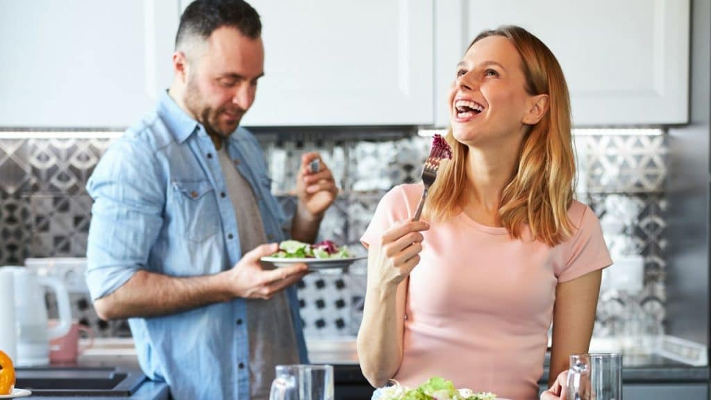 A woman laughs while eating salad in a kitchen as a man stands behind her holding food.