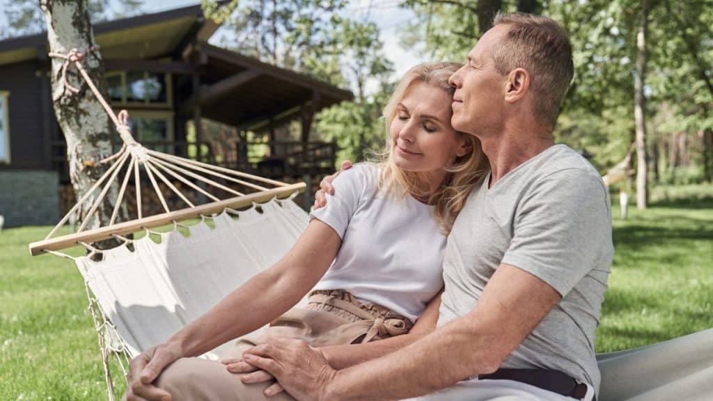 A man and woman sit in a white hammock outdoors with a house in the background.