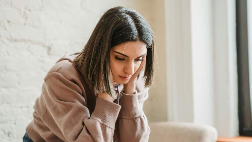 A woman in a brown hoodie sits with her head resting in her hands.