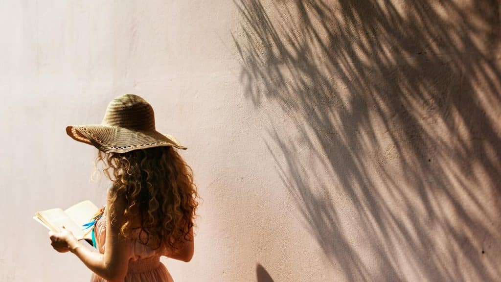 A person wearing a sun hat reads a book beside a sunlit wall.