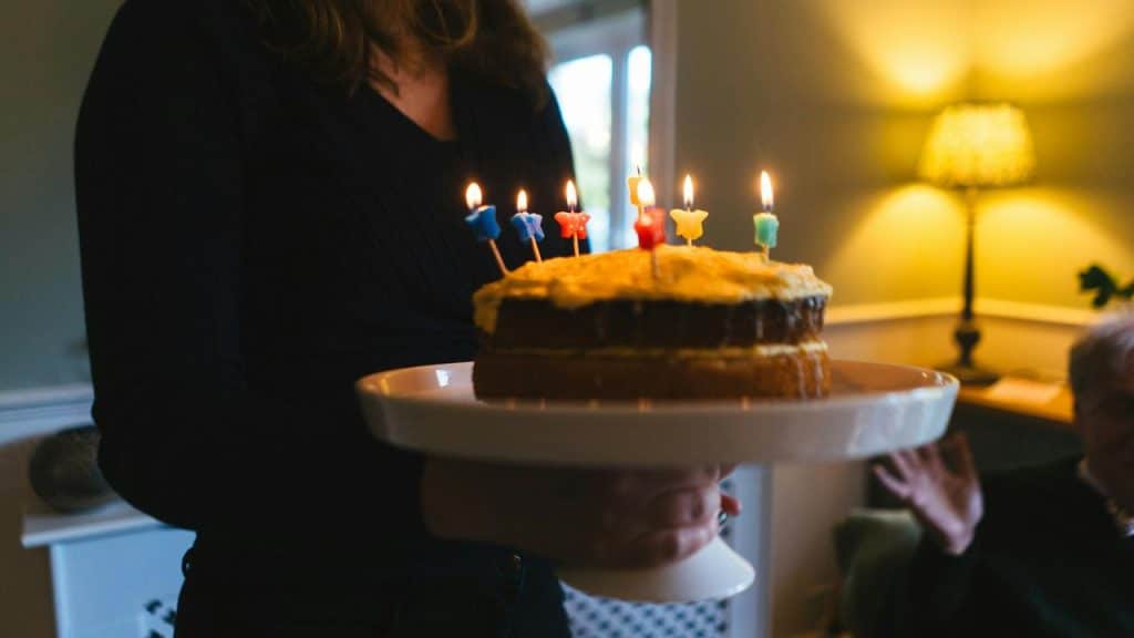 A person holds a birthday cake with lit candles in a warmly lit room.