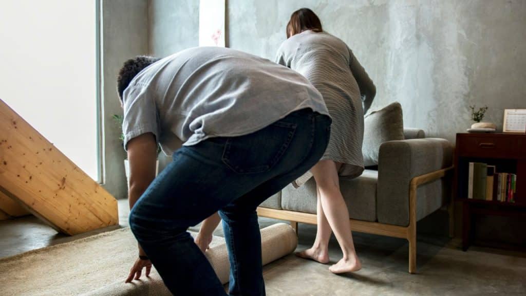 Two people in a room are bending over to roll up a large beige rug together.