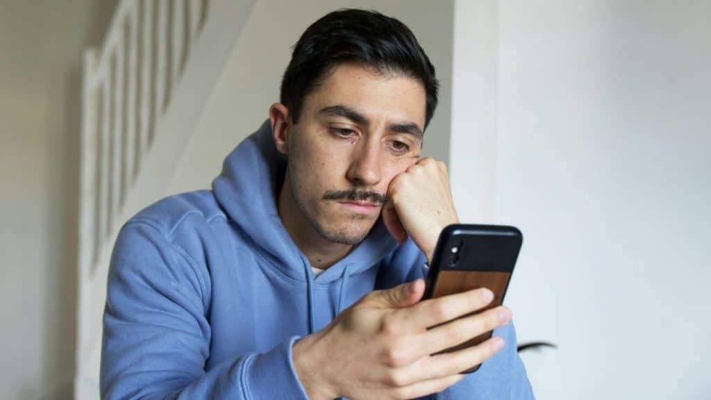 A man with a mustache wearing a blue hoodie looks sadly at his smartphone.