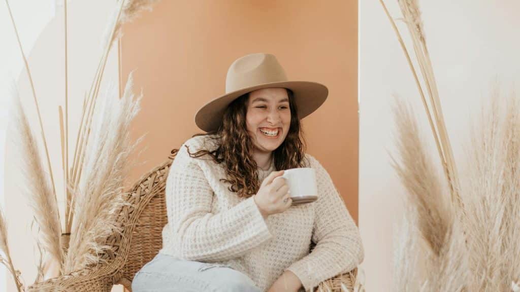 A woman in a wide-brim hat smiling while holding a mug in a cozy setting.