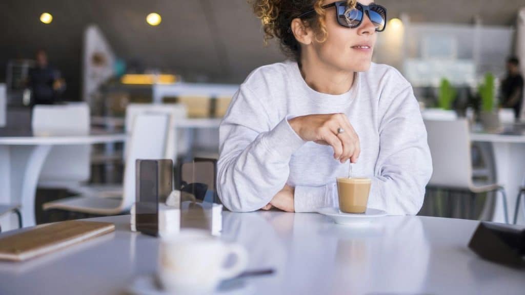 A woman wearing sunglasses sits at a café table with a cup of coffee.