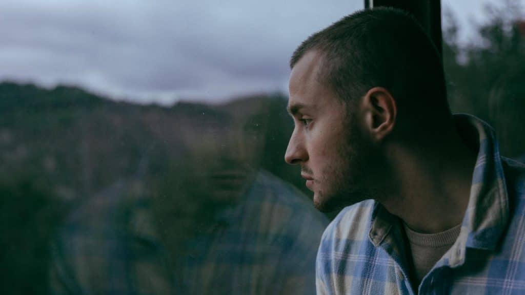 A man in a plaid shirt looks out a window with his reflection visible in glass.