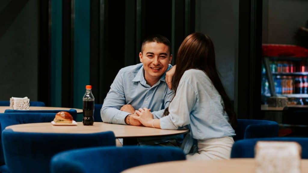 A smiling man and a woman sit at a round table holding hands in a restaurant.