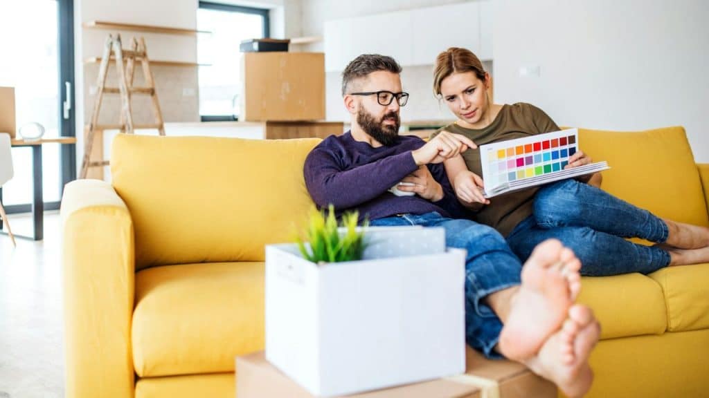 A man and woman sit on a yellow sofa looking at a color swatch book.