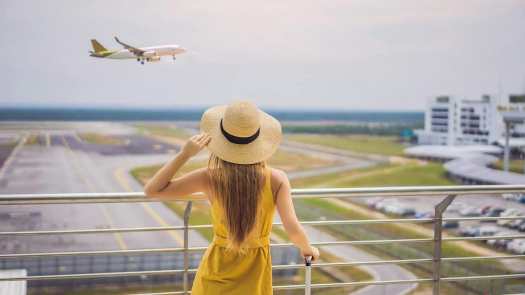 A woman is adjusting her hat and holding a bag while looking at a plane take off from a balcony by the airport.