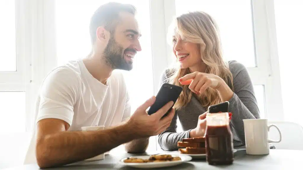 A couple laughs while looking at a phone during breakfast.