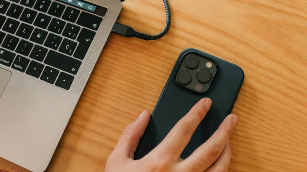 A hand holding a smartphone next to a laptop on a wooden desk.