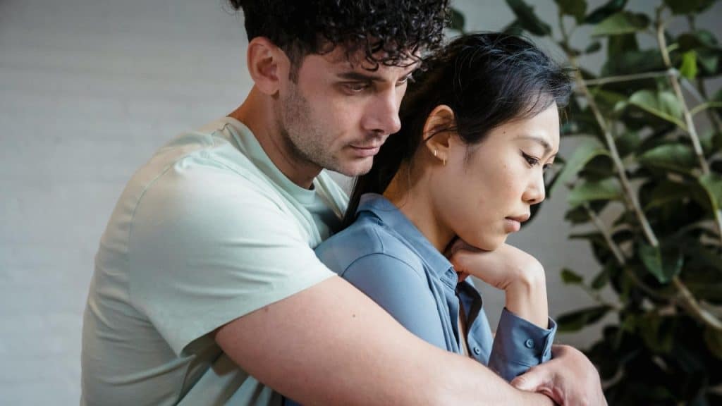 A man with curly hair hugs a woman from behind as she looks down thoughtfully.