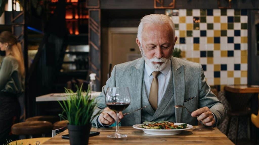 A senior man in a gray suit eating a meal alone at a restaurant table.