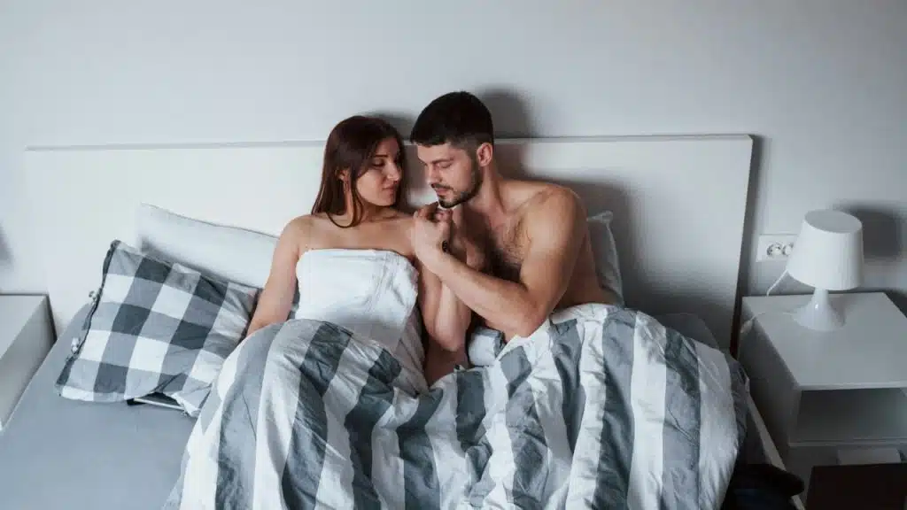 A man and woman sit together in a bed, holding hands and looking at each other.