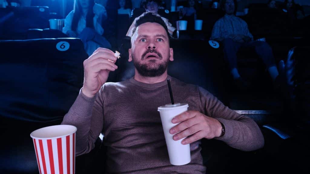 A man in a theater seat looks forward with wide eyes, holding popcorn and drink.