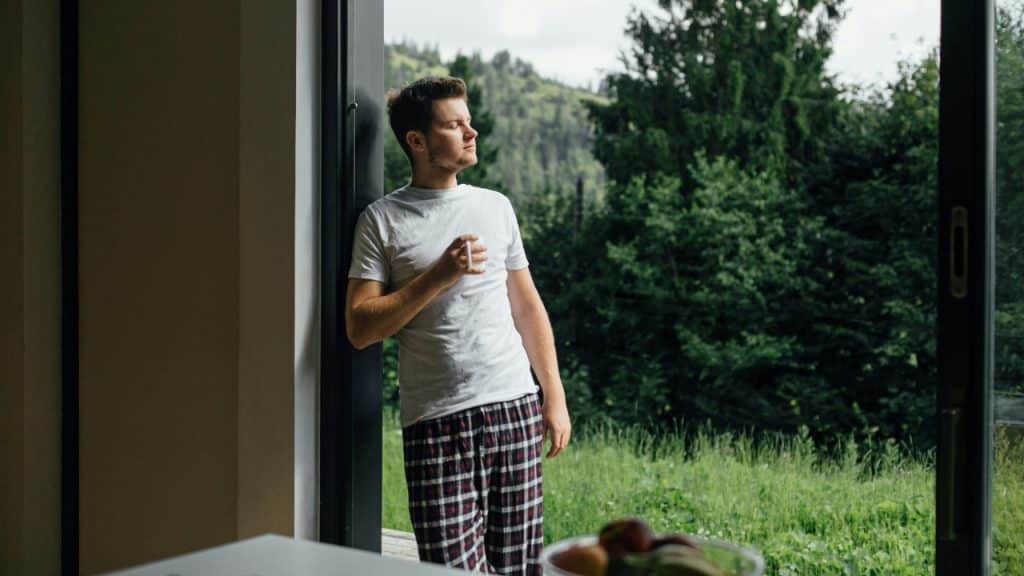 A man in pajamas stands in a doorway holding a mug and looking at trees.
