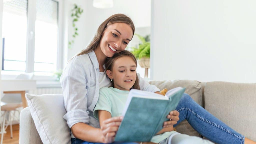 A woman and a young girl sit together on a couch reading a blue book.